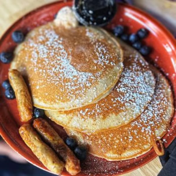Just Brunch pancakes with powdered sugar, blueberries, sausage links, and syrup on a red plate.