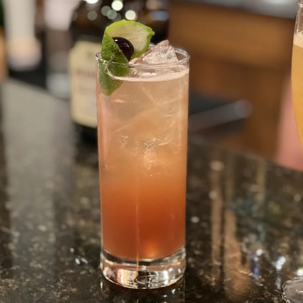 A tall glass of iced brunch cocktail, garnished with a lime peel and a cherry, on a dark counter.