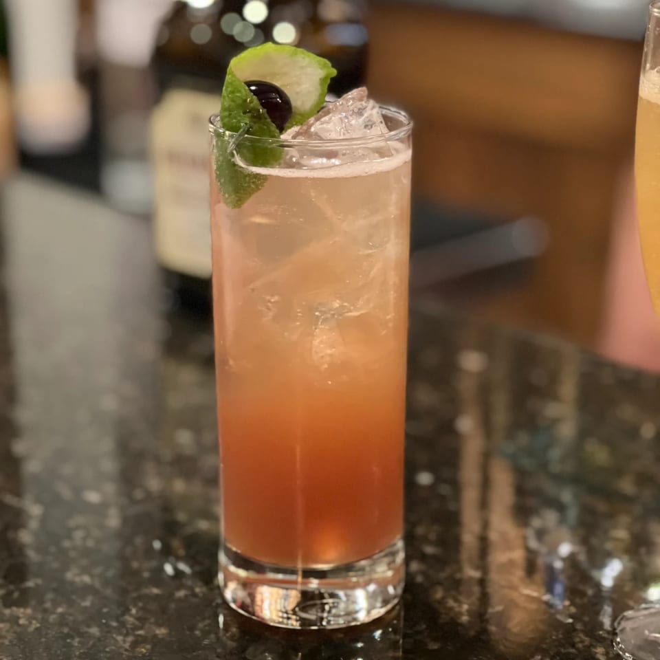 A tall glass of iced brunch cocktail, garnished with a lime peel and a cherry, on a dark counter.