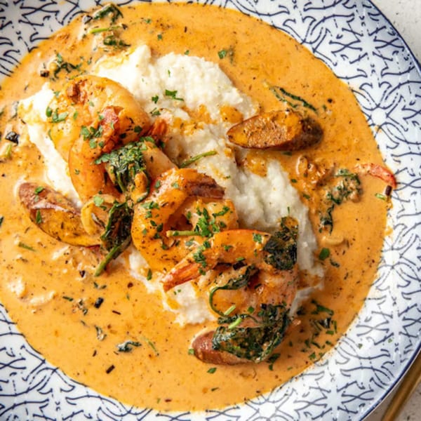 Shrimp and grits with a creamy orange sauce, sausage, and greens on a decorative plate.
