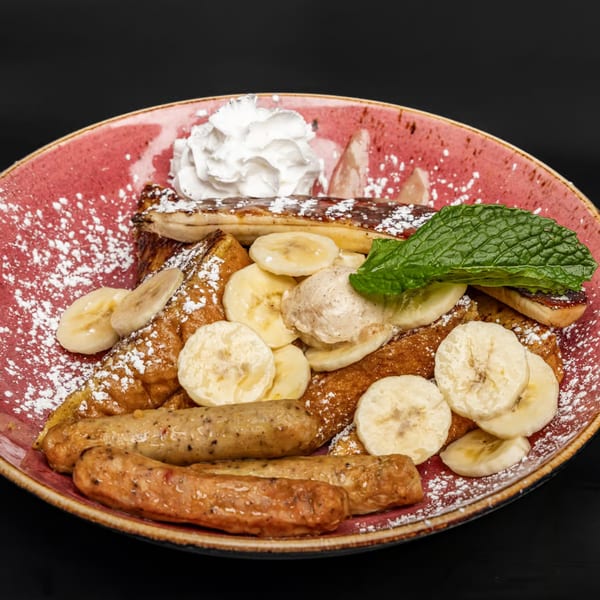 Just Brunch's French toast with bananas, whipped cream, sausage, and powdered sugar on a red plate.