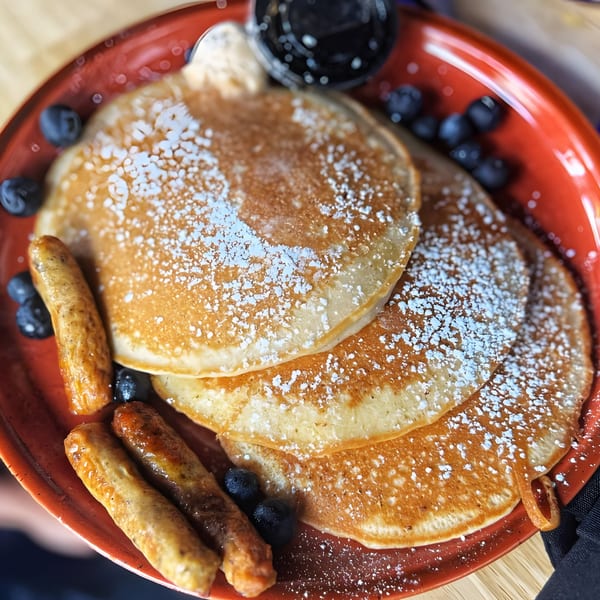 Just Brunch pancakes with powdered sugar, blueberries, sausage links, and syrup on a red plate.