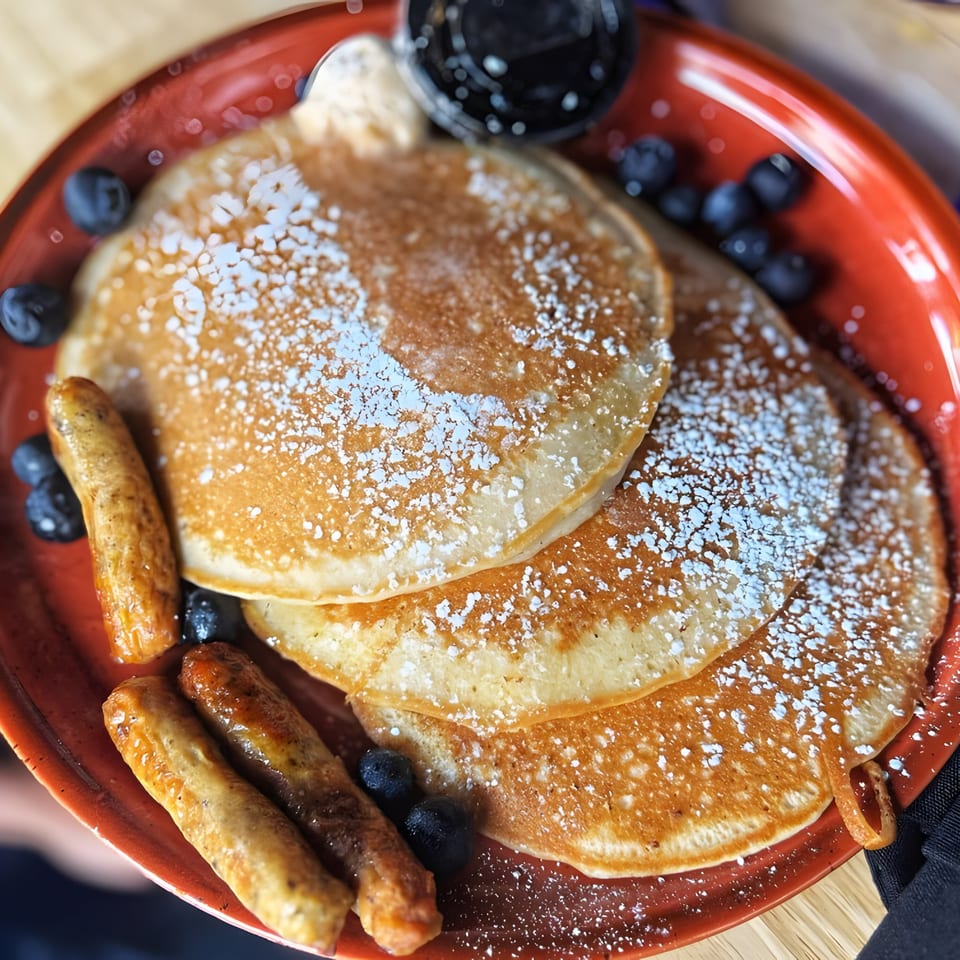 Just Brunch pancakes with powdered sugar, blueberries, sausage links, and syrup on a red plate.