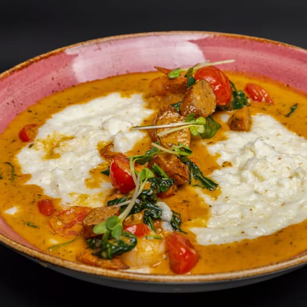 Just Brunch's shrimp and grits with cherry tomatoes, spinach, and microgreens in a pink bowl.