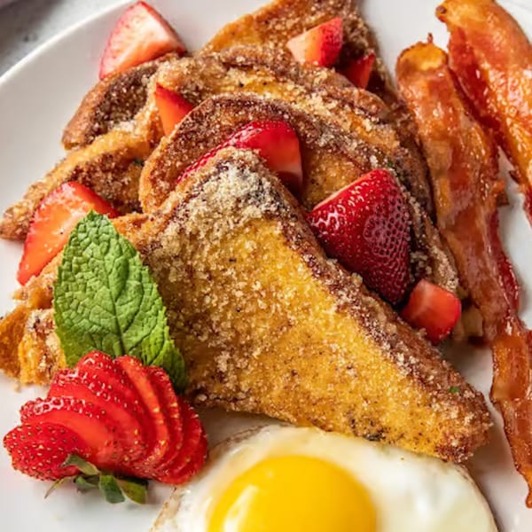 Just Brunch's French toast with strawberries, bacon, a fried egg, and mint on a white plate.
